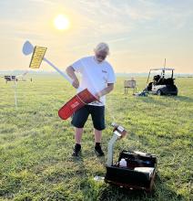 Man assembling red and white model plane on grassy field at sunrise.