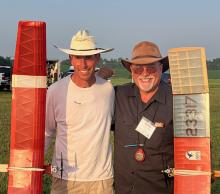 Two men smiling, wearing hats, stand outdoors holding model planes.