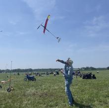 Man launching model glider in grassy field under clear blue sky.