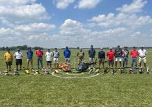 Group of people standing on grass with model helicopters under a blue sky.