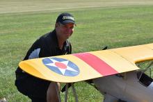 Man smiling next to a model airplane with a yellow wing and emblem on grass.