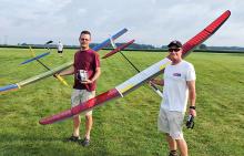 Two men holding colorful model gliders on a grassy field.