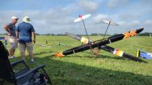 Two men stand near model gliders on a grassy field under a cloudy sky.