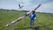 Man holding large model glider on grassy field under blue sky.