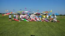 Group of people posing with colorful model gliders on a grassy field.