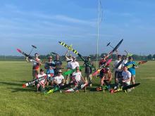 Group of people holding model aircraft on a grassy field.