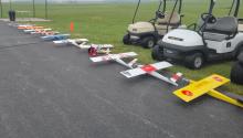 Model airplanes lined up on a paved road near golf carts and grassy fields.