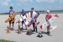 Four people holding model airplanes, standing with trophies on a tarmac.
