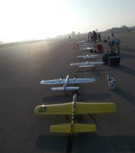 Row of model airplanes on a runway at sunrise, with people prepping nearby.