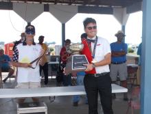 Young man holding a trophy, smiling; other people present in a pavilion setting.