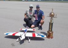 Two people kneel by a large trophy and model airplane on a tarmac.