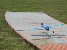 A small blue model plane taking off from a wooden runway on grass.