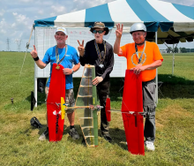 Three men posing with model airplanes under a tent on a grassy field.