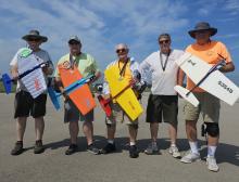Five men with remote control planes, smiling, standing on pavement under clear sky.