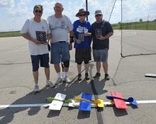 Four men holding plaques stand beside colorful model airplanes on pavement.