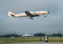 RC plane flying low over a field, man controlling it on the ground.
