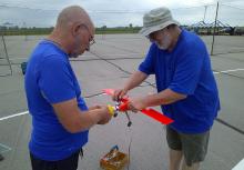 Two men in blue shirts fixing a red model airplane outdoors.