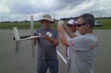 Three men on an airfield inspect a model rocket, holding fuel container and tubing.