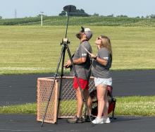 Two people watch the sky with a camera on a tripod in a grassy field.