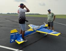 Two men standing by a large blue and yellow model plane on asphalt.