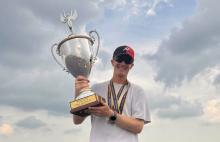 Man smiling, holding a large trophy outdoors, wearing a red and black cap.