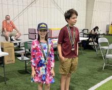 Two children wearing medals stand on artificial turf indoors, smiling.