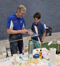 Man and boy building a model airplane together at a workshop table.