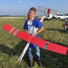 Young boy holding a large red model airplane outdoors on a grassy field.