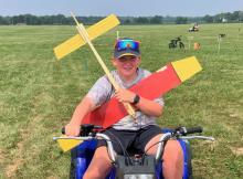 Boy smiles on an ATV holding red and yellow model plane on grassy field.