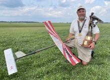 Man kneeling in a field holding a large trophy and an RC glider, smiling, wearing a medal.