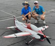 Two men crouching beside a large remote control airplane on a parking lot.