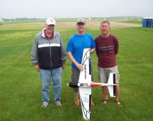Three men standing on grass with a model airplane.
