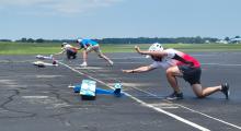 Three people launching model planes on a runway, wearing helmets.