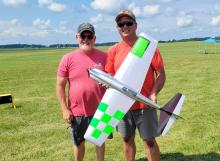 Two men smiling, holding a model airplane on a grassy field.