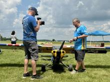 Man films another near small model plane on grass field.
