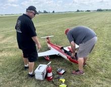 Two men at a grassy field inspecting a red model aircraft.