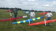 Three men holding colorful model gliders on grassy field.