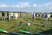 Model gliders on grass with a group of people in a field, talking and observing.