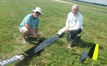 Two men kneeling beside a large black remote-control plane on grass.
