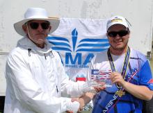 Two men shaking hands; one holds a trophy. A logo banner in the background.