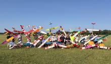Group of people holding colorful model gliders in a grassy field.